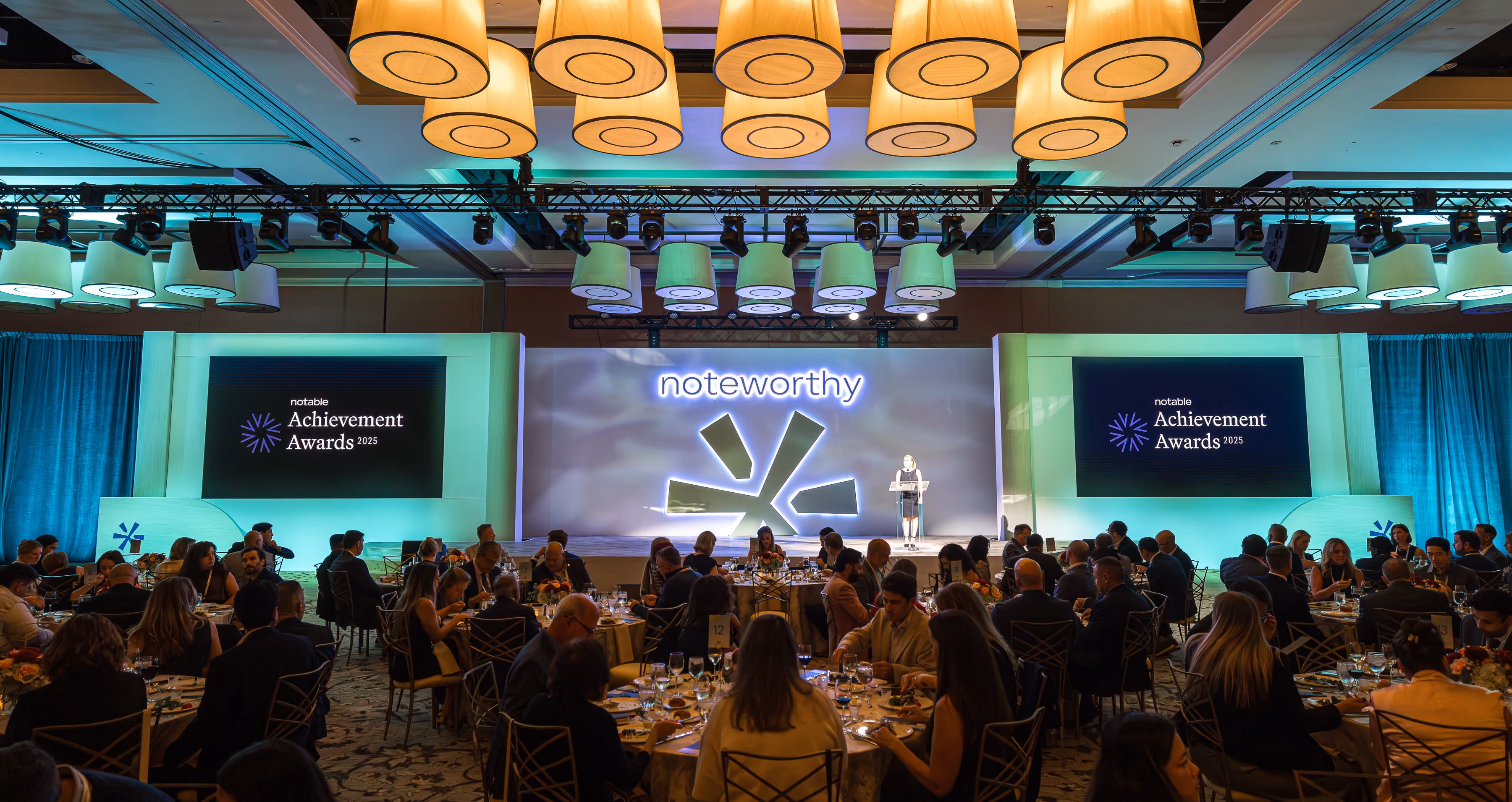 Audience seated at round tables with a speaker on stage at a Noteworthy Achievement Awards event.