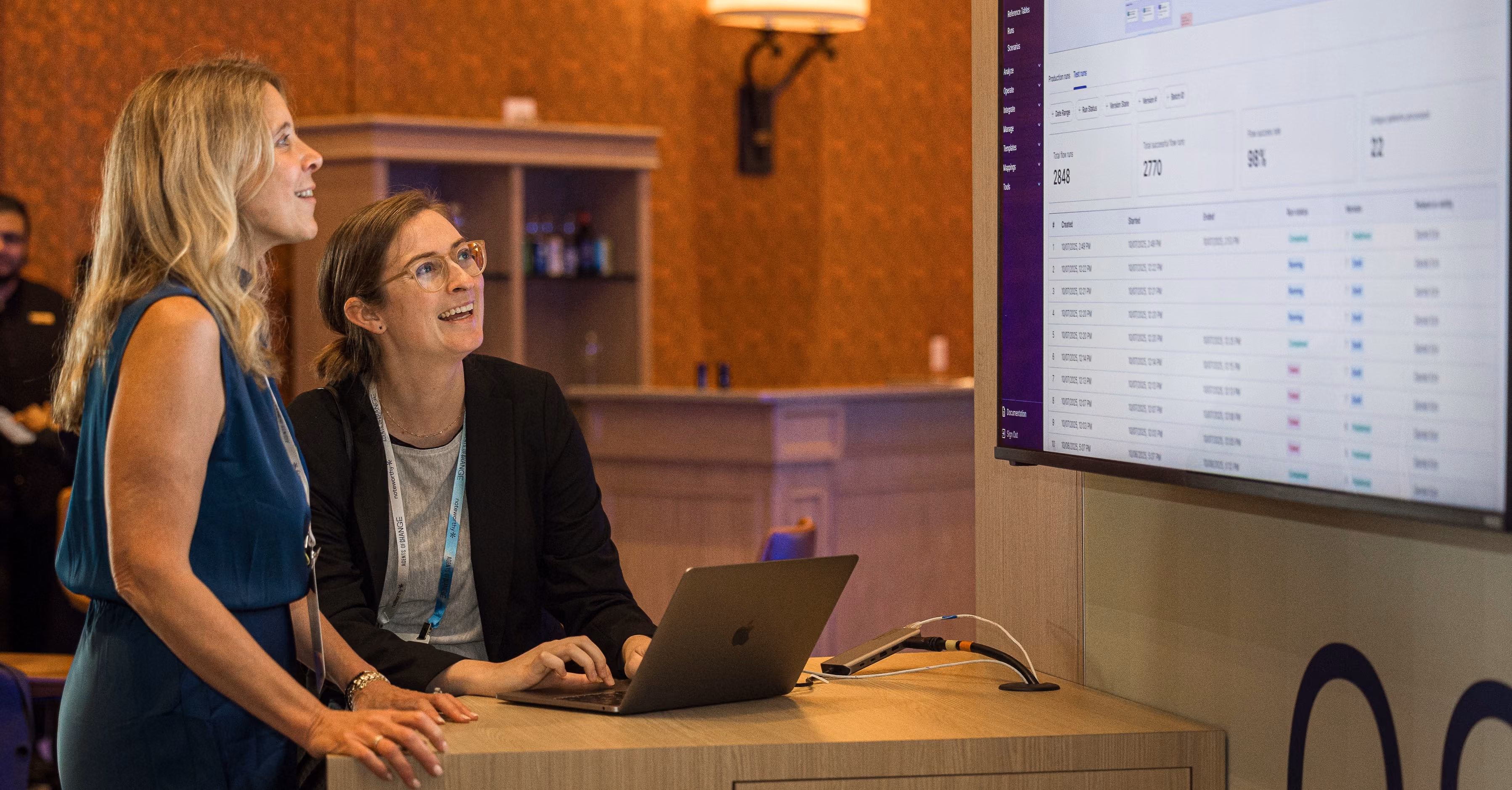 Two women smiling and looking at a large screen displaying data, one typing on a laptop.