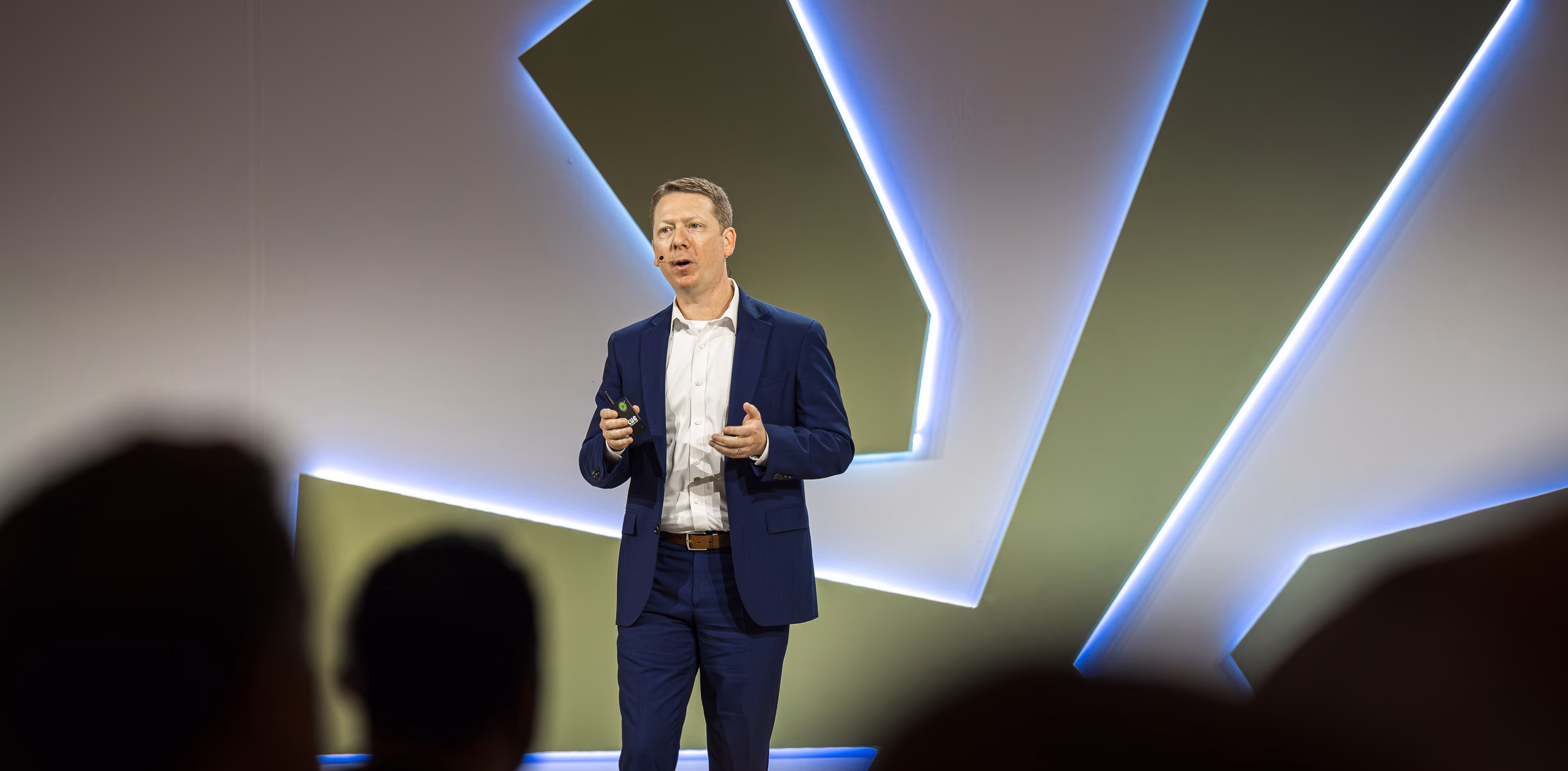 Man in blue suit speaks with headset microphone on stage with modern geometric lighted backdrop.