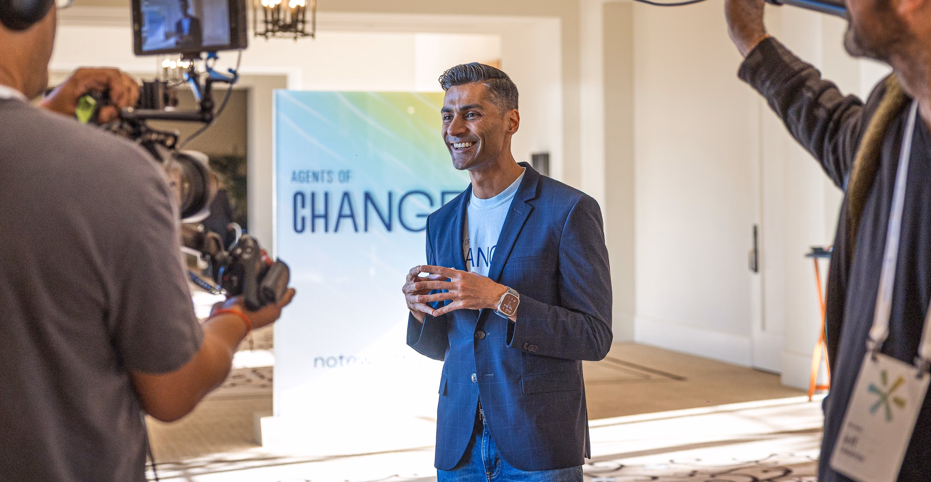 Smiling man in blazer speaking on camera in a conference room with Agents of Change sign behind him.