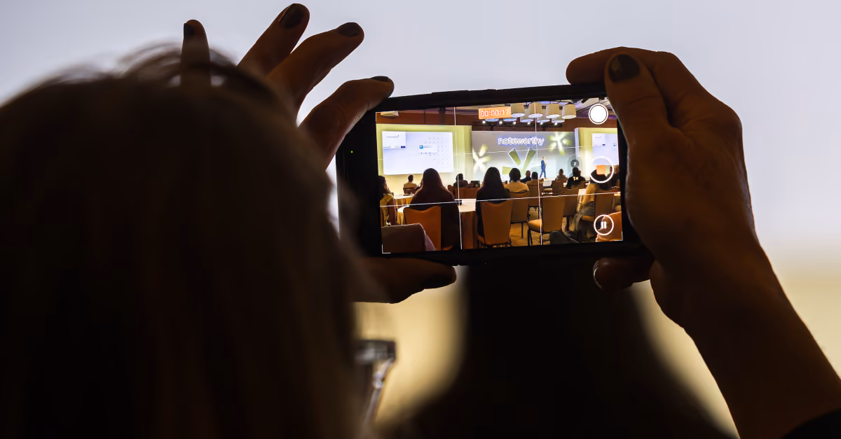Person recording a speaker presenting on a stage at an indoor conference with a smartphone.