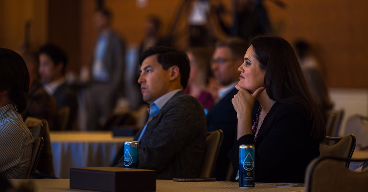 Two attentive adults seated at a conference table with blue Proud Source beverage cans in front of them.
