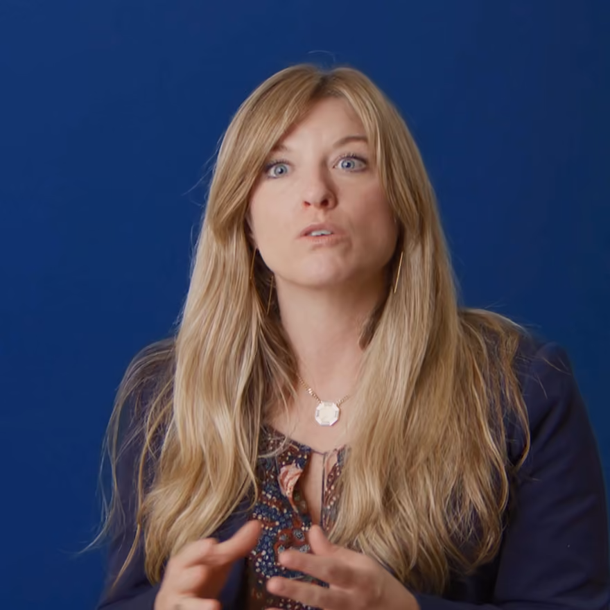 Woman with long blonde hair speaking, wearing a patterned blouse and dark blazer against a blue background.