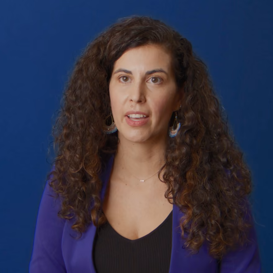 Woman with long curly hair wearing a purple blazer and black top against a dark blue background.