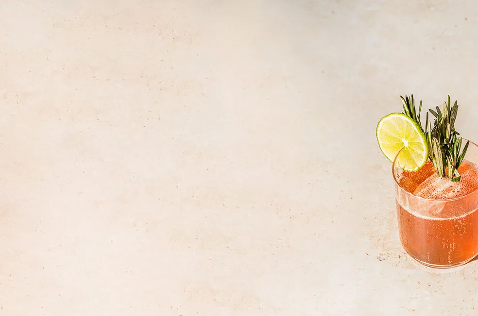 ein roter Drink mit Rosmarinzweig und Limettenscheibe auf beige-farbenem Hintergrund