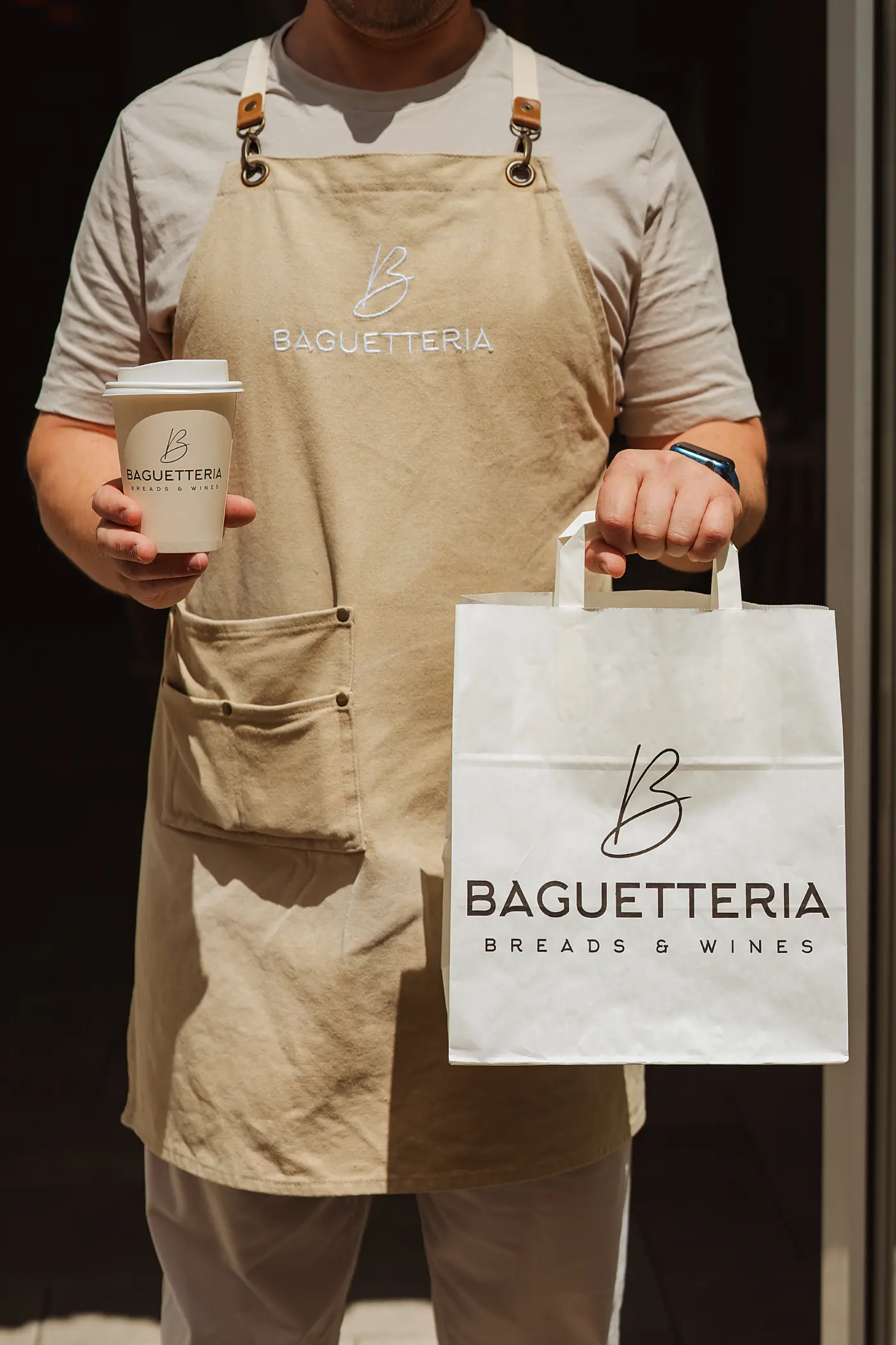 A person wearing a beige apron with Baguetteria logo holds a paper coffee cup and a white paper bag, both branded with Baguetteria Breads & Wines logo.