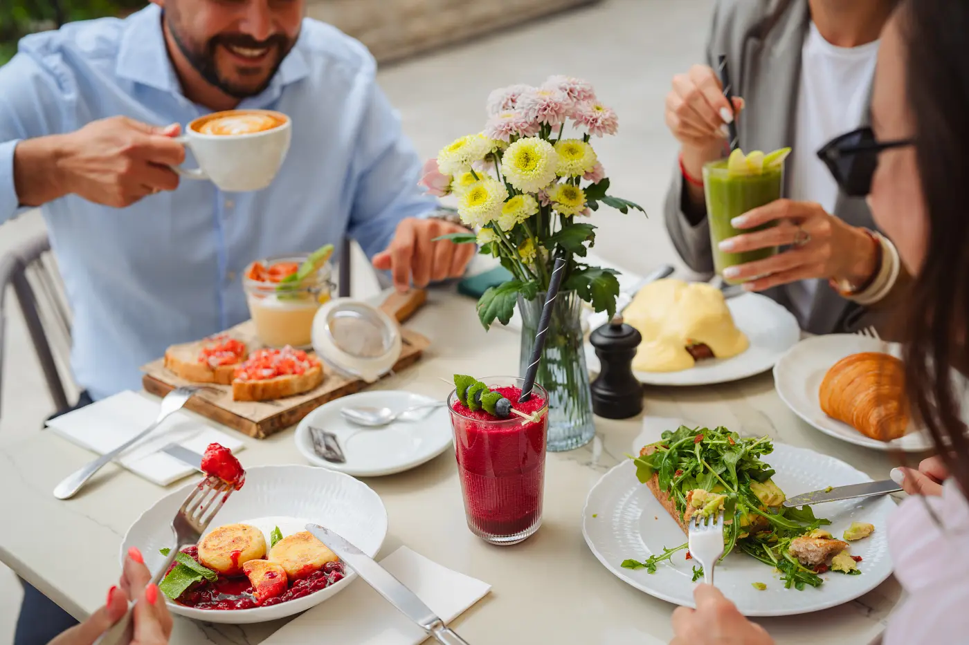 Three people enjoy a brunch at an outdoor table set with pastries, omelets, salad, a berry smoothie, and coffee, with a vase of flowers in the center.