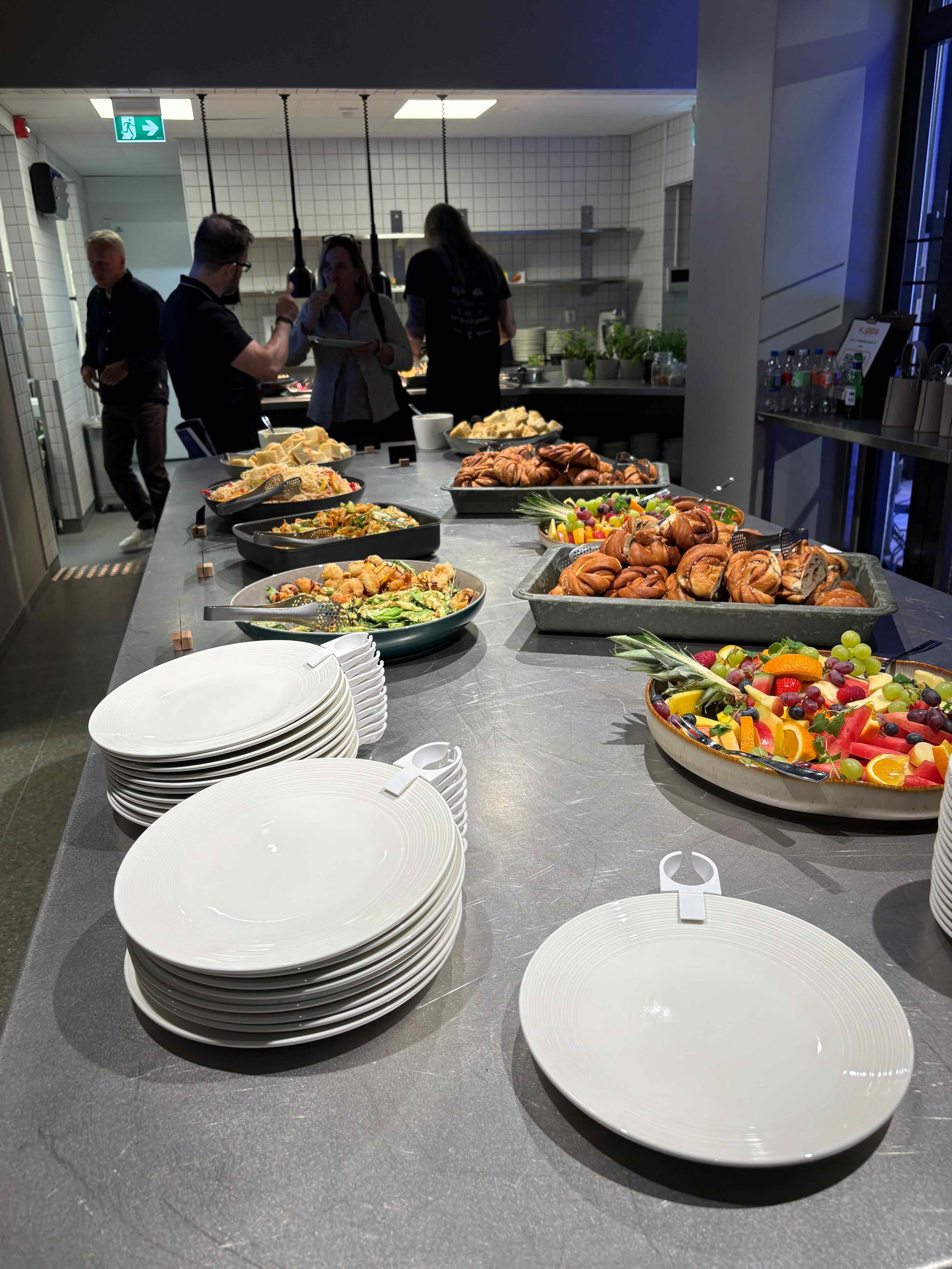 A long buffet table covered with food. At the front are tall stacks of white plates, and behind them are platters of salads, cinnamon rolls, fruit platters, and bottled water. In the background, several people are helping themselves at an open kitchen with white tiles.