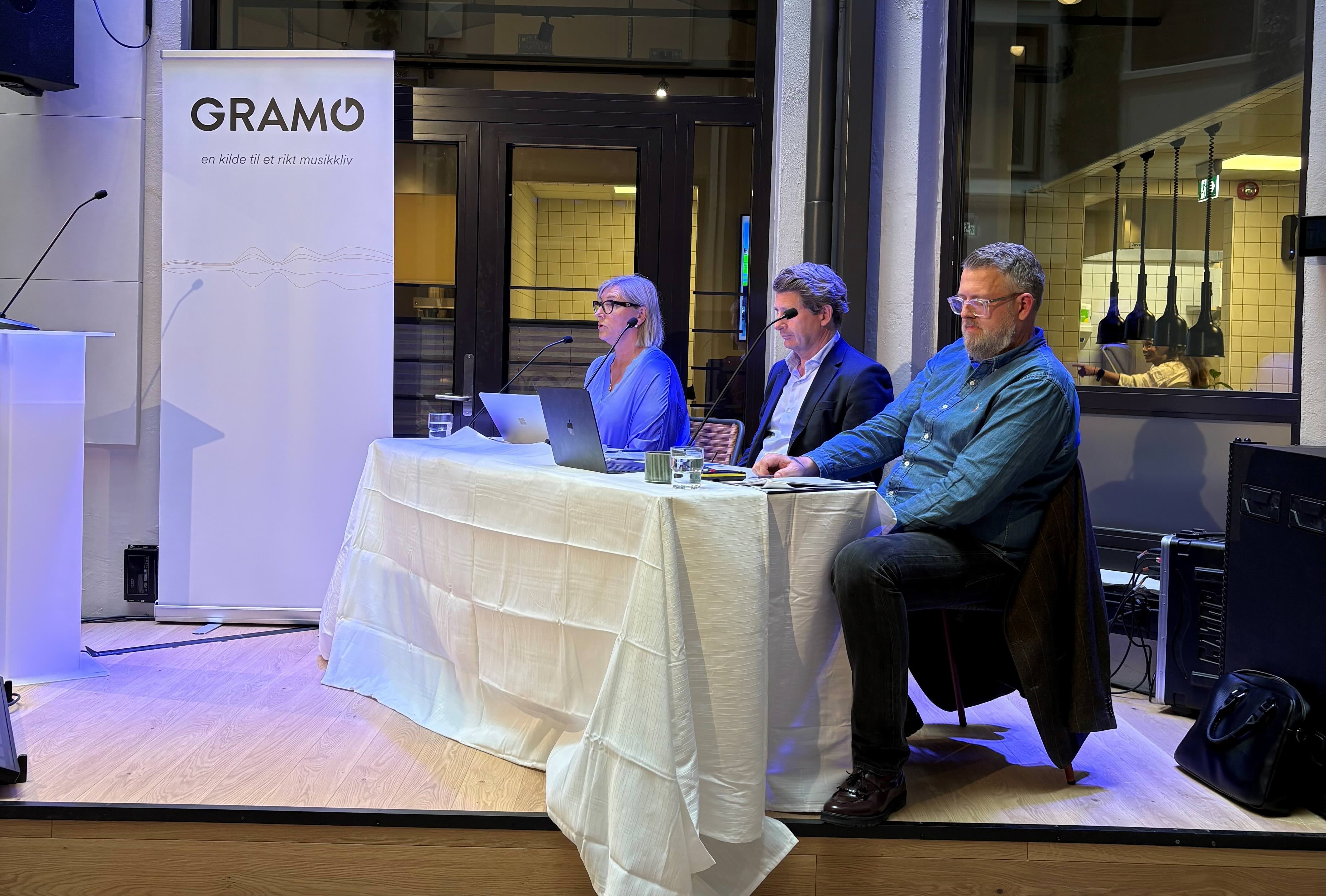 Three people are sitting on stage during Gramo's annual meeting. Behind them, Gramo's logo hangs on the wall, and in front of the stage is a low table with a white tablecloth and a sign marked "Gramo."