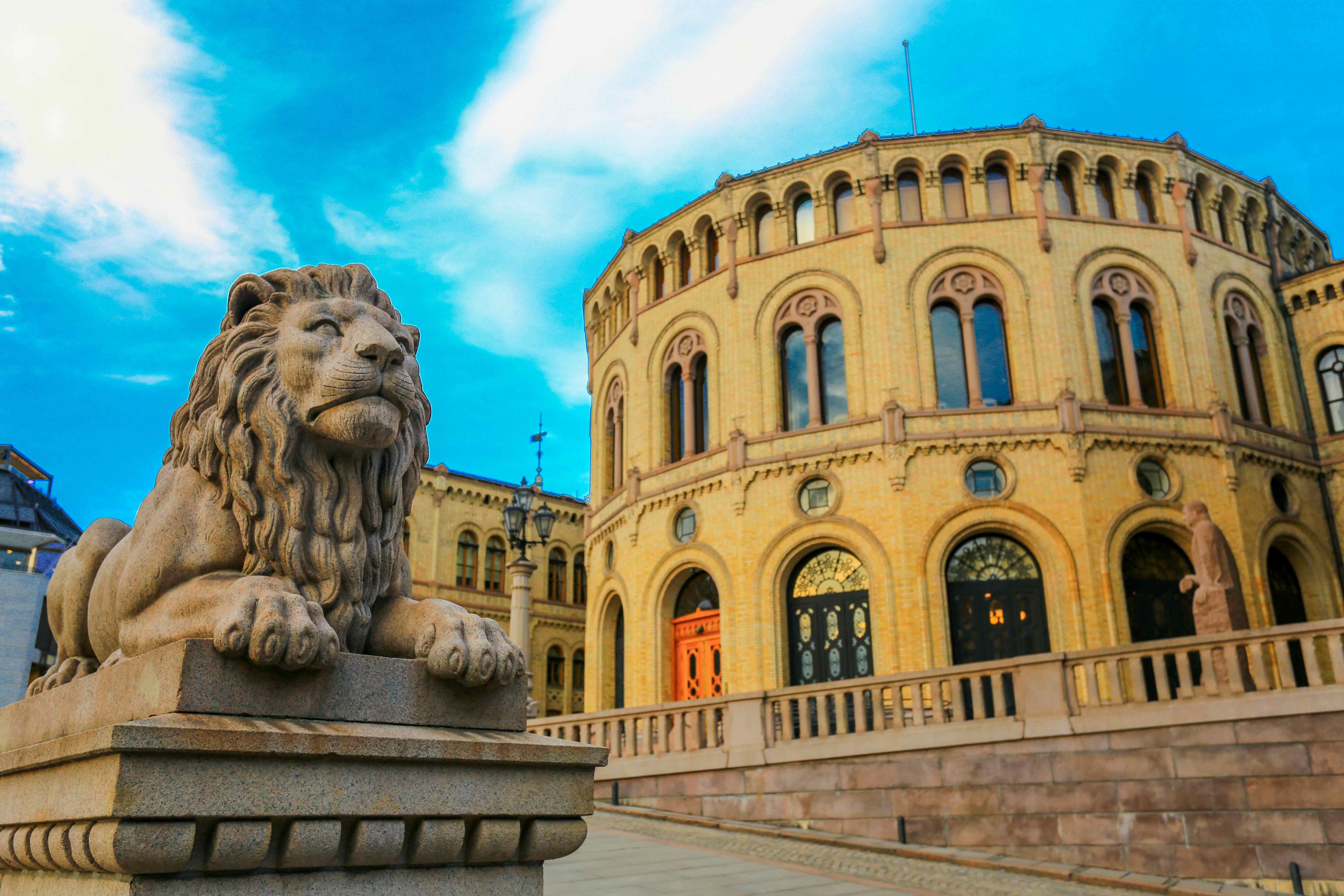 One of the lion sculptures in front of the Storting in the foreground, with the curved yellow brick facade and entrance to the Storting building behind. The photo is taken from below against a blue sky with clouds.