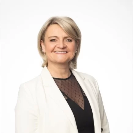 Smiling woman with blonde hair wearing a white blazer over a black top with sheer detail.