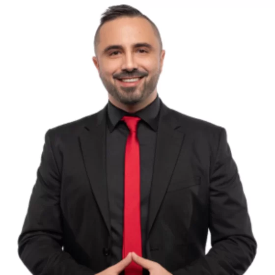 Smiling man in a black suit and red tie with hands clasped in front against white background.