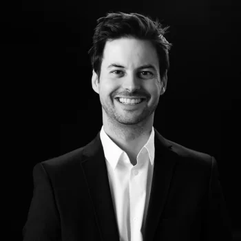Smiling man with dark hair wearing a black blazer and white shirt against a black background.