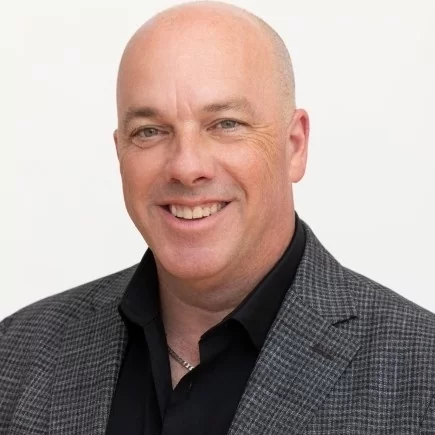 Bald man in a gray checkered jacket and black shirt smiling against a white background.