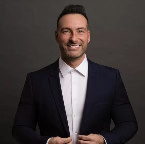 Smiling man in dark suit and white shirt adjusting jacket against a dark background.