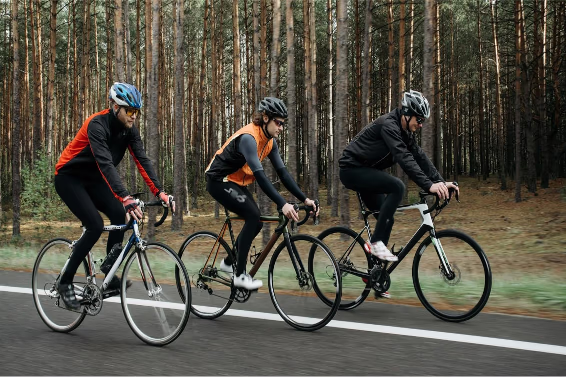 Three cyclists wearing helmets and athletic gear riding road bikes on a forested road.
