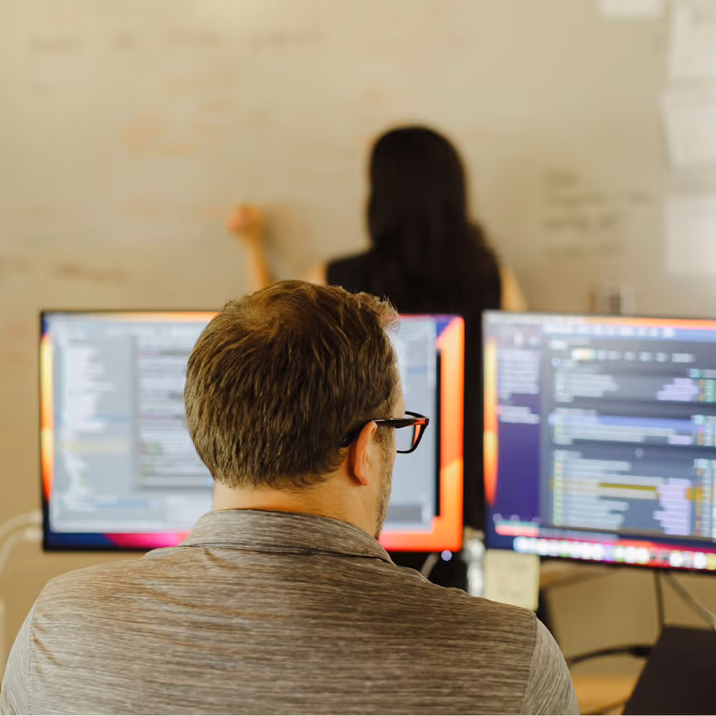 A person works at a desk with dual monitors, while another writes on a whiteboard, collaborating on AI solutions.
