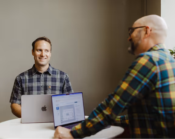 Two men wearing plaid shirts sitting at a round table using laptops and engaged in conversation.