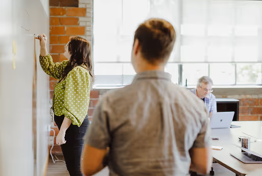 Woman writing on a whiteboard in a meeting room with two men, one seated at a desk with a laptop and the other standing with his back to the camera.