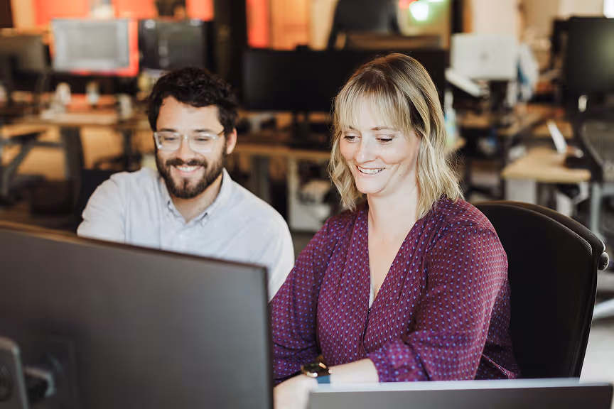 Two coworkers smiling and looking at a computer screen in a modern office setting.