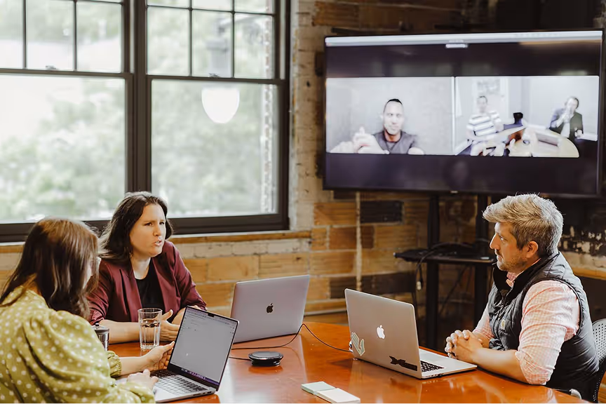 Three people in a meeting room with laptops, participating in a video conference displayed on a large screen.