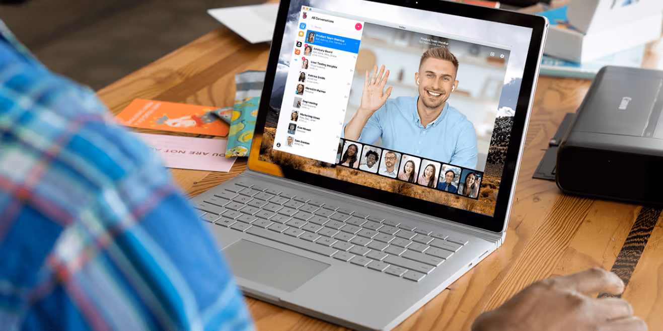 Person in a blue plaid shirt participating in a video call on a laptop showing a man waving and multiple participants.