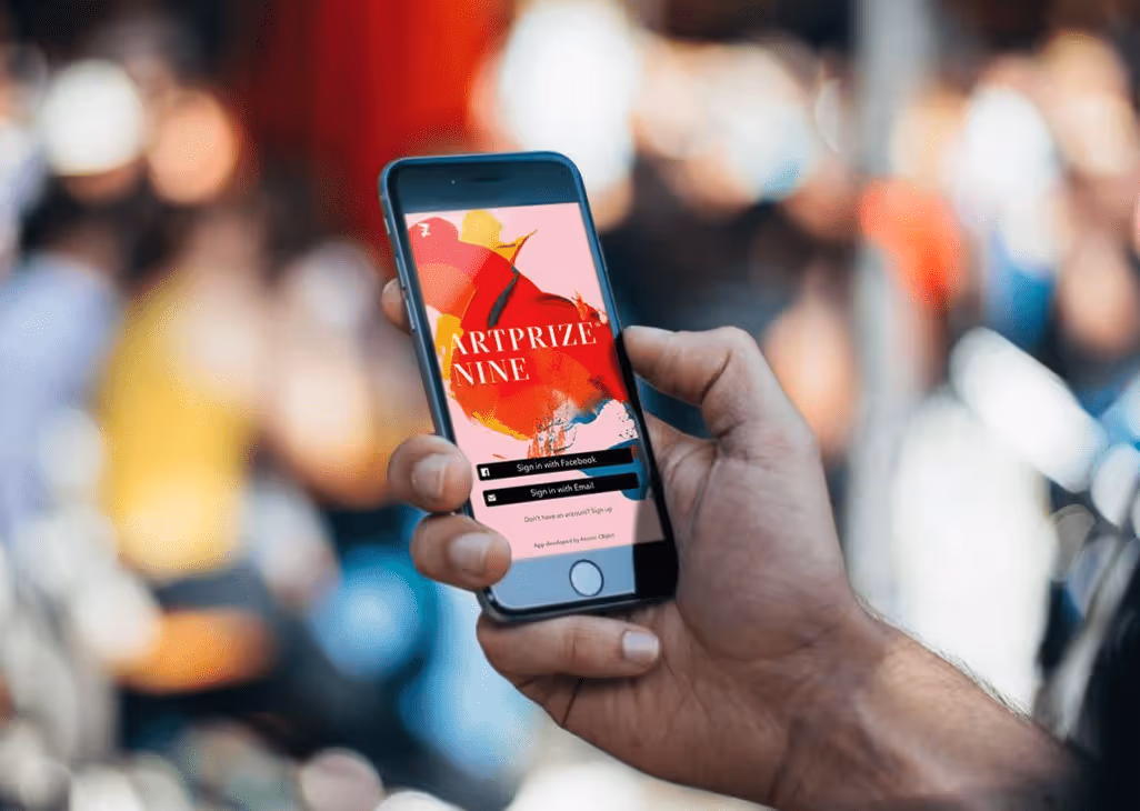 Hand holding smartphone displaying ArtPrize Nine app login screen with options to sign in via Facebook or Email.