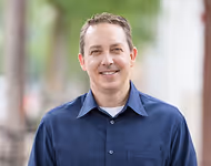 Smiling man wearing a dark blue button-up shirt standing outdoors with blurred greenery and building in the background.