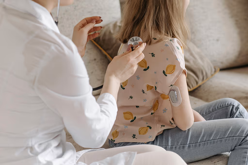 Medical professional using a stethoscope to listen to the back of a young girl with a medical patch on her arm.