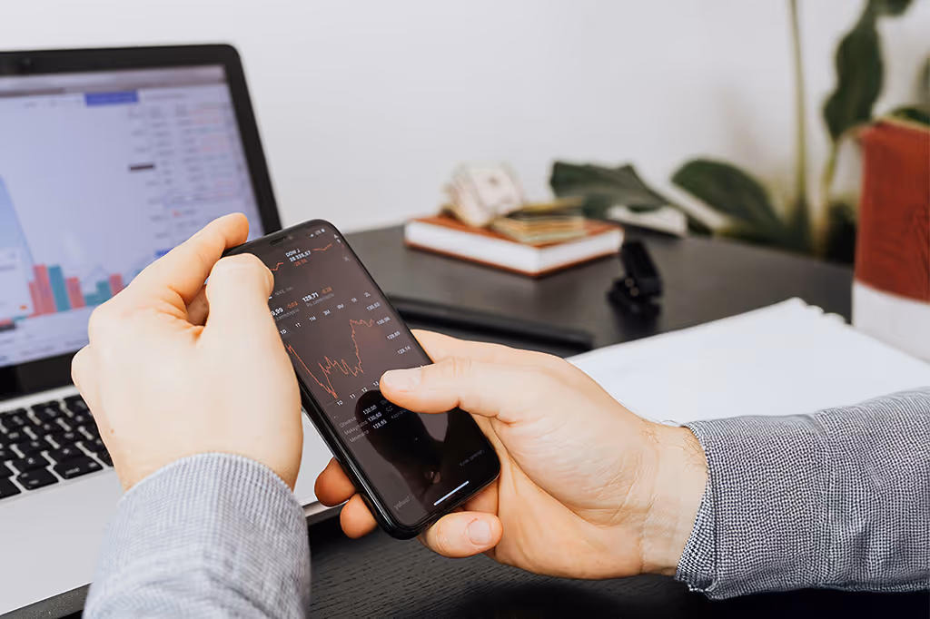 Person holding a smartphone displaying financial stock charts with a laptop showing graphs in the background.