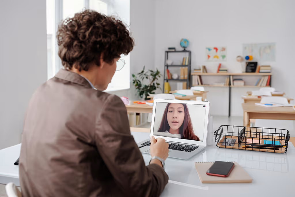 Person with curly hair and glasses sitting at a desk, engaging in a video call with a young girl displayed on a laptop screen in a classroom setting.