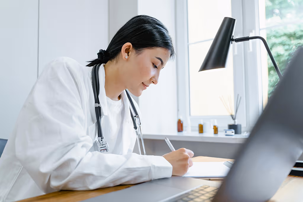 Female doctor with a stethoscope around her neck writing notes at a desk in a bright office.