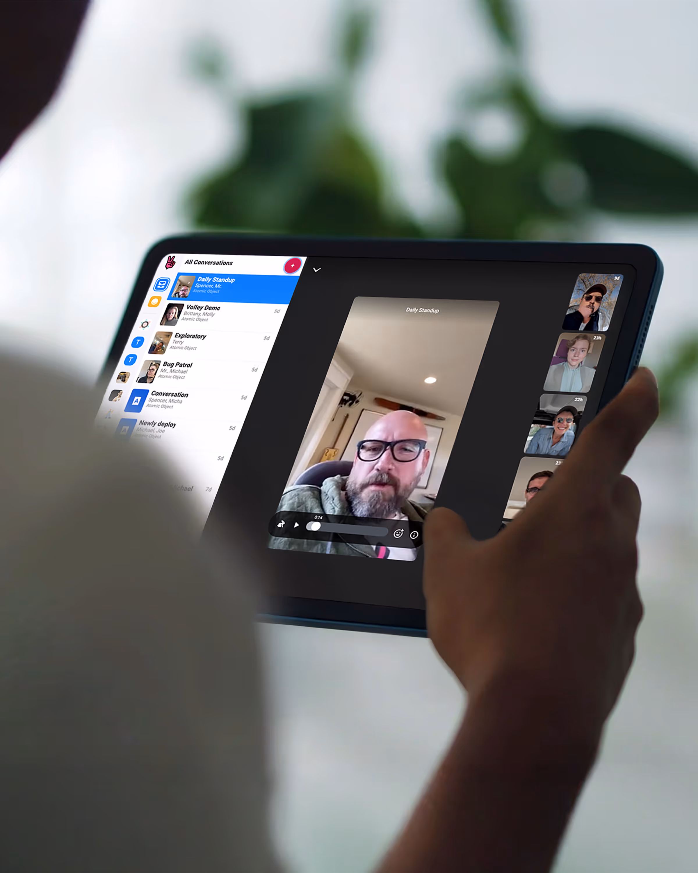 Person holding a tablet displaying a video call with multiple participants and a list of conversation threads on the side.