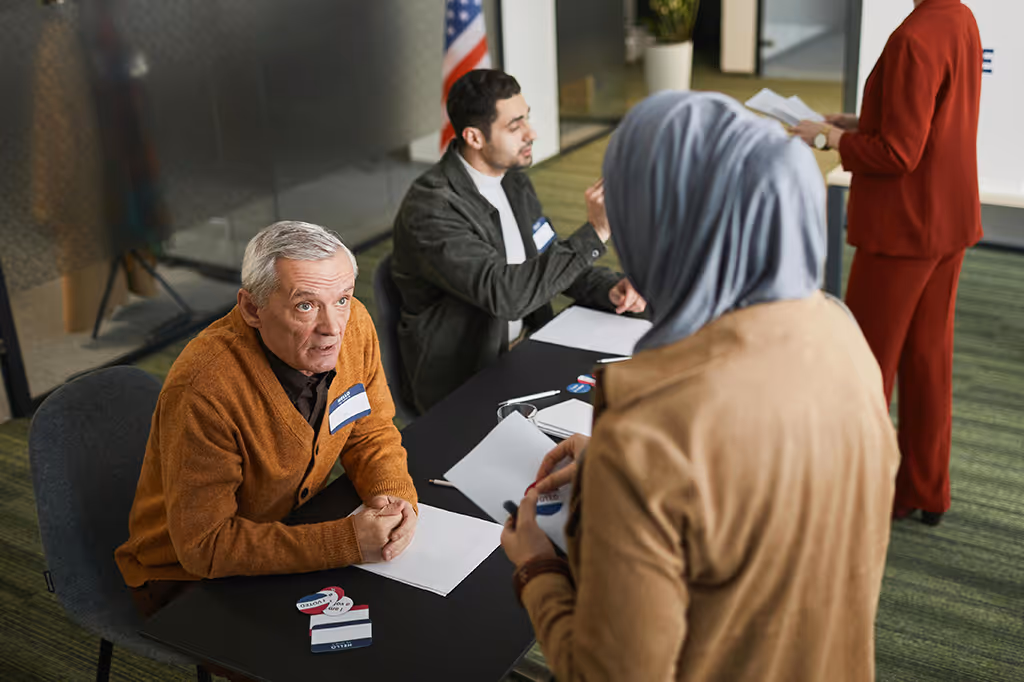 An elderly man and a younger man seated at a table with papers and voting stickers, interacting with a woman wearing a hijab at a voting or registration event indoors.