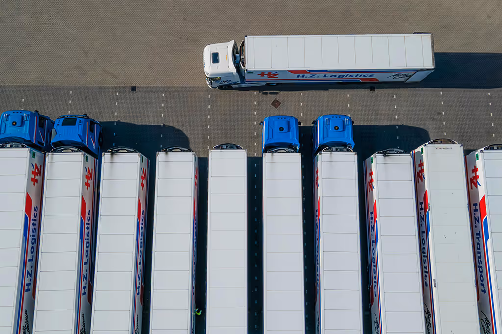 Aerial view of multiple white semi-trucks with blue cabs lined up in a parking area labeled H.Z. Logistics and H.Z. Transport.