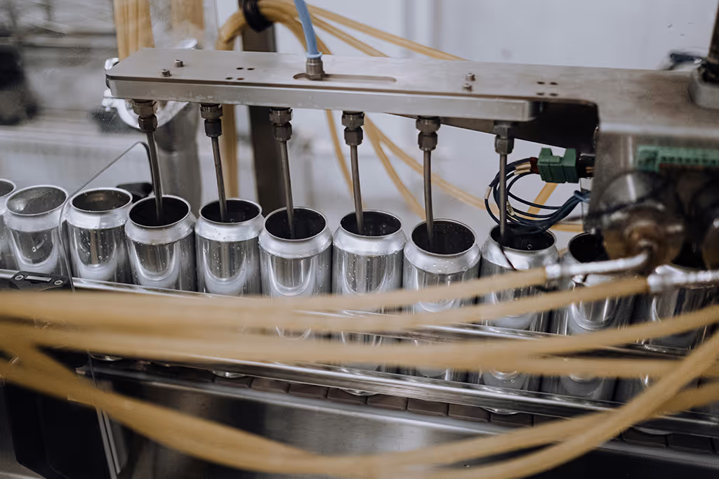 Industrial machine filling rows of empty aluminum cans with liquid in a manufacturing setting.
