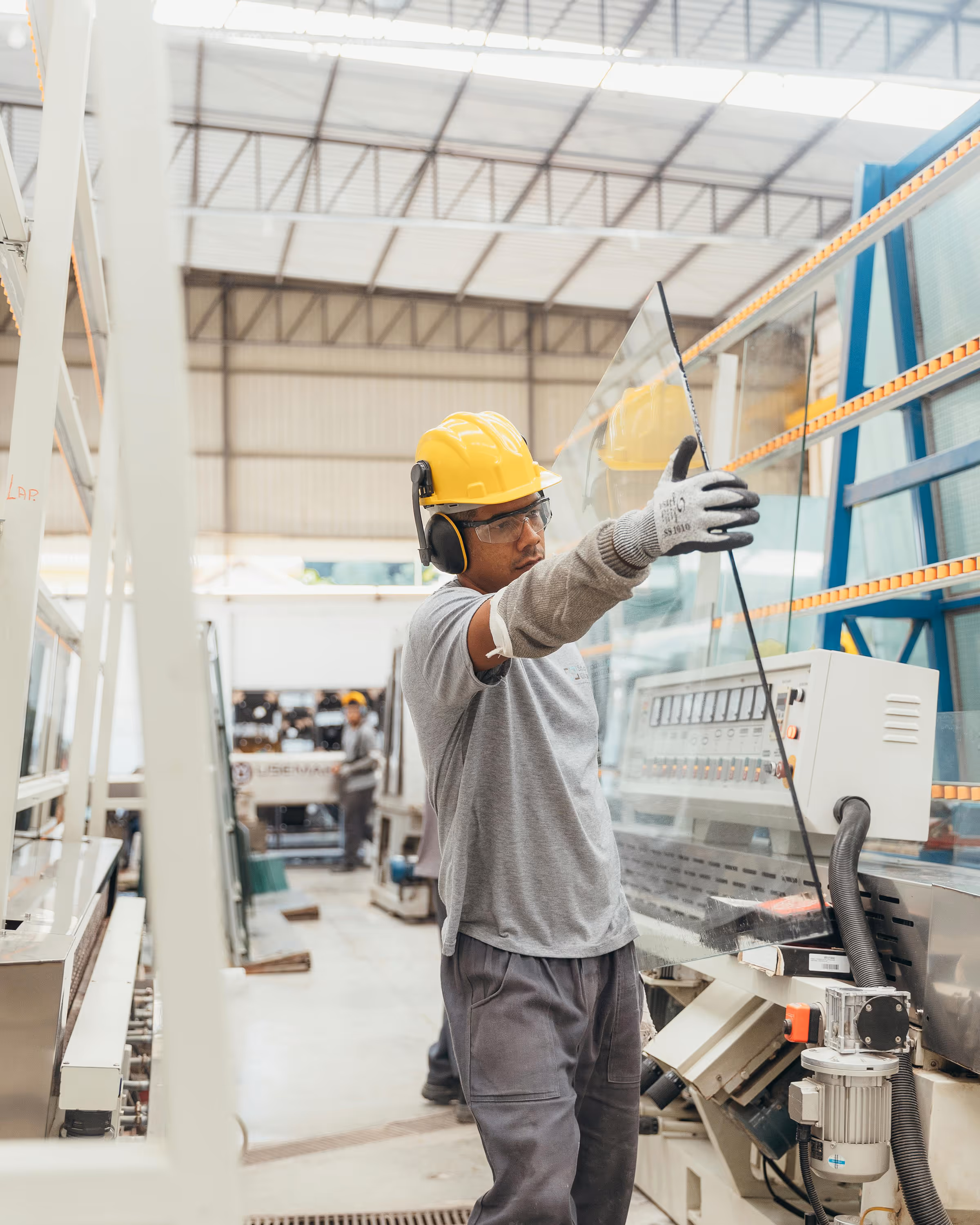 Worker wearing safety gear handling a large glass panel in a manufacturing facility.