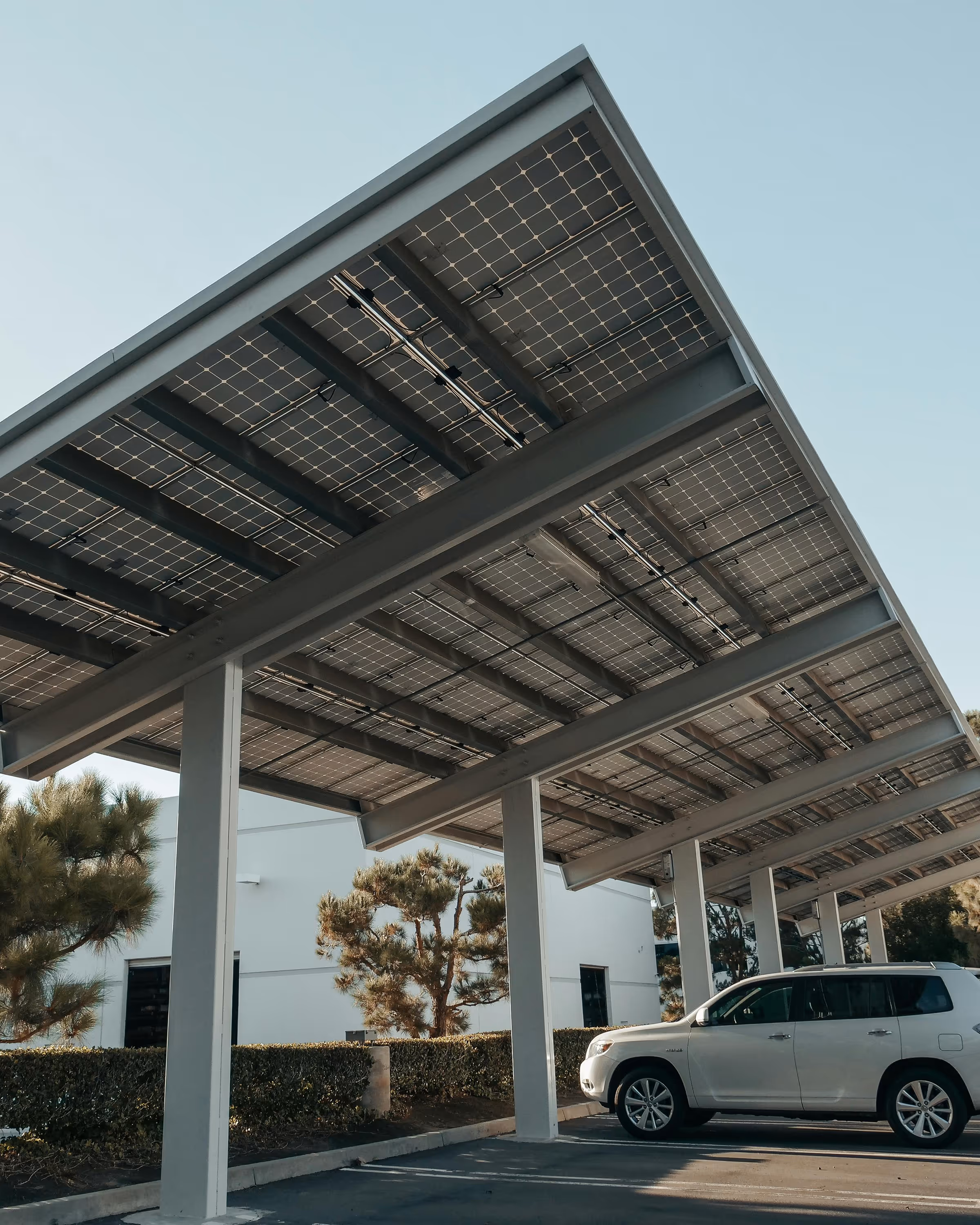 Electric car parked under a solar panel carport with trees and a white building in the background.