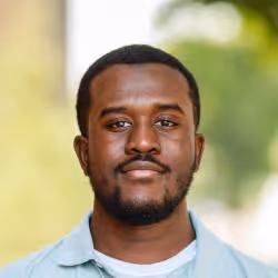 Portrait of a man with short hair and beard wearing a light blue shirt against a blurred nature background.