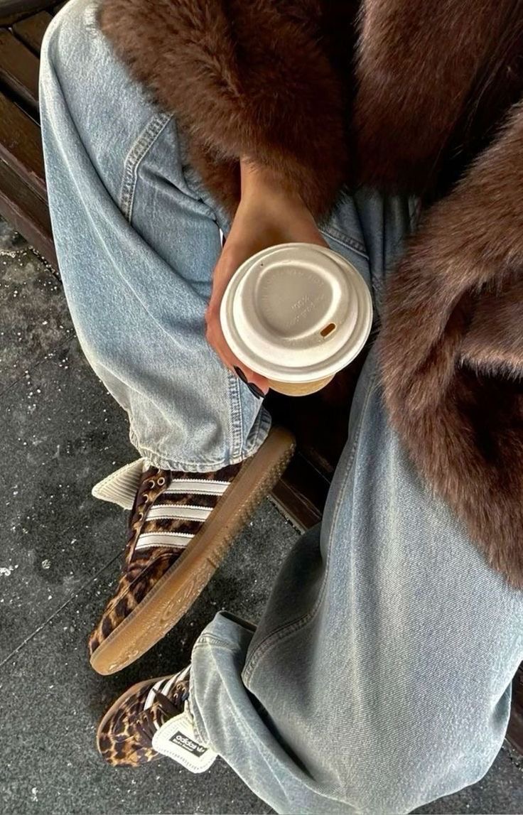 Close up of a girl in jeans and a fur coat paired with leopard print Adidas Samba sneakers and a cup of coffee.
