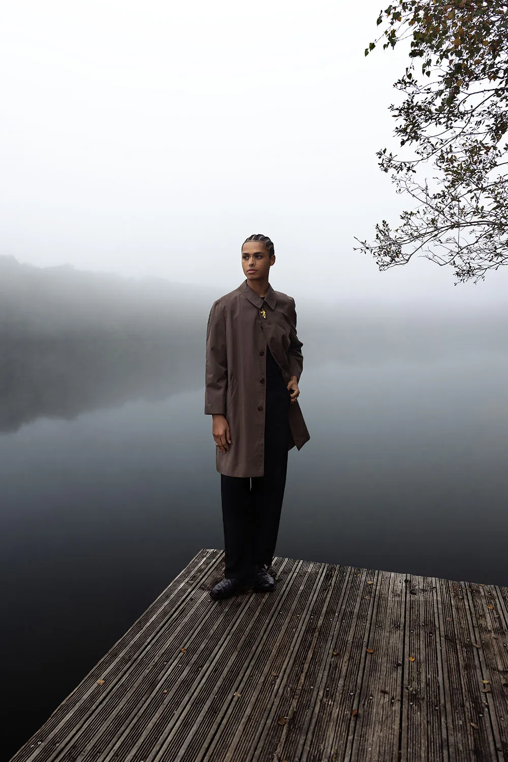Shooting éditorial masculin sur un ponton, mannequin debout face à une lac brumeux, créant une atmosphère mystérieuse.