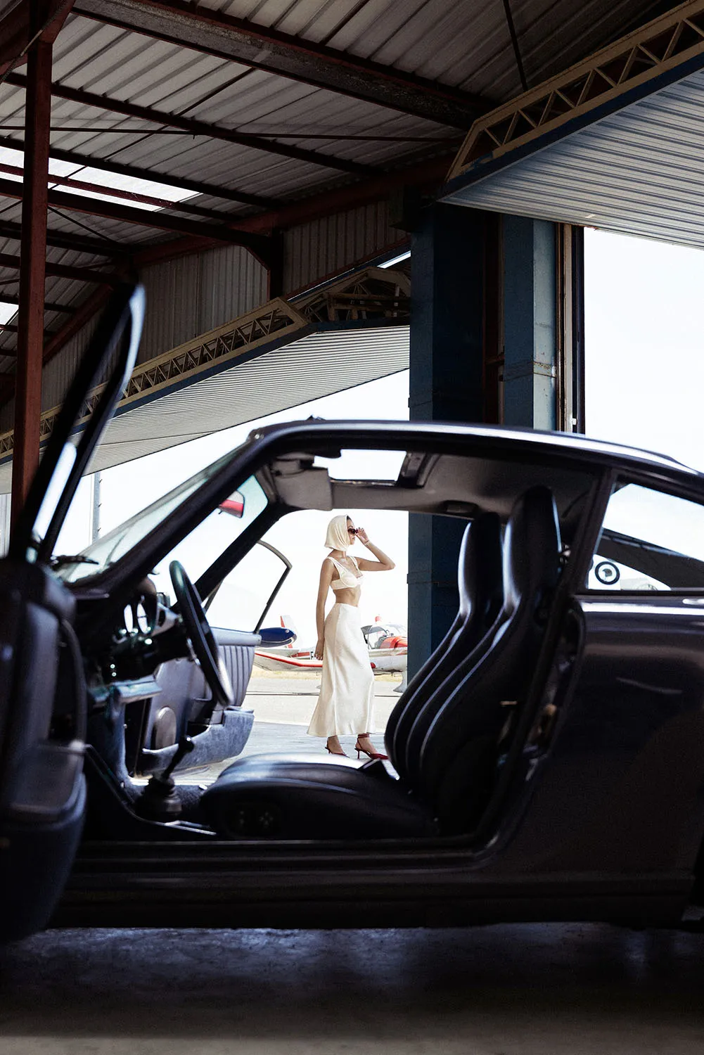 Jeu de perspective de plans, shooting fashion et éditorial avec une Porsche 911 bleu foncé. On voit une mannequin à travers les portes ouvertes