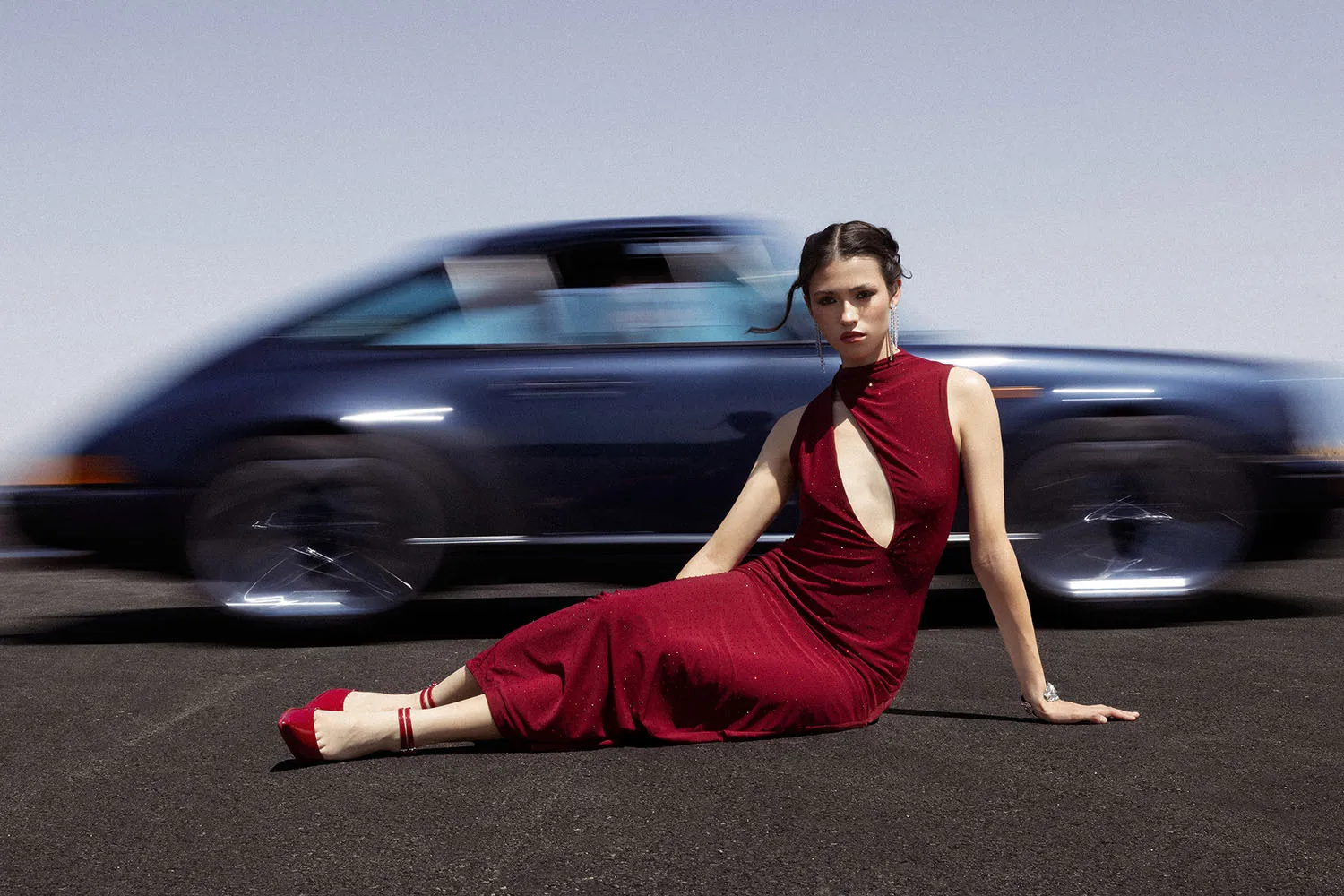 Shooting fashion et éditorial avec une Porsche 911 bleu. Une mannequin en robe rouge pose devant la voiture en mouvement de flou