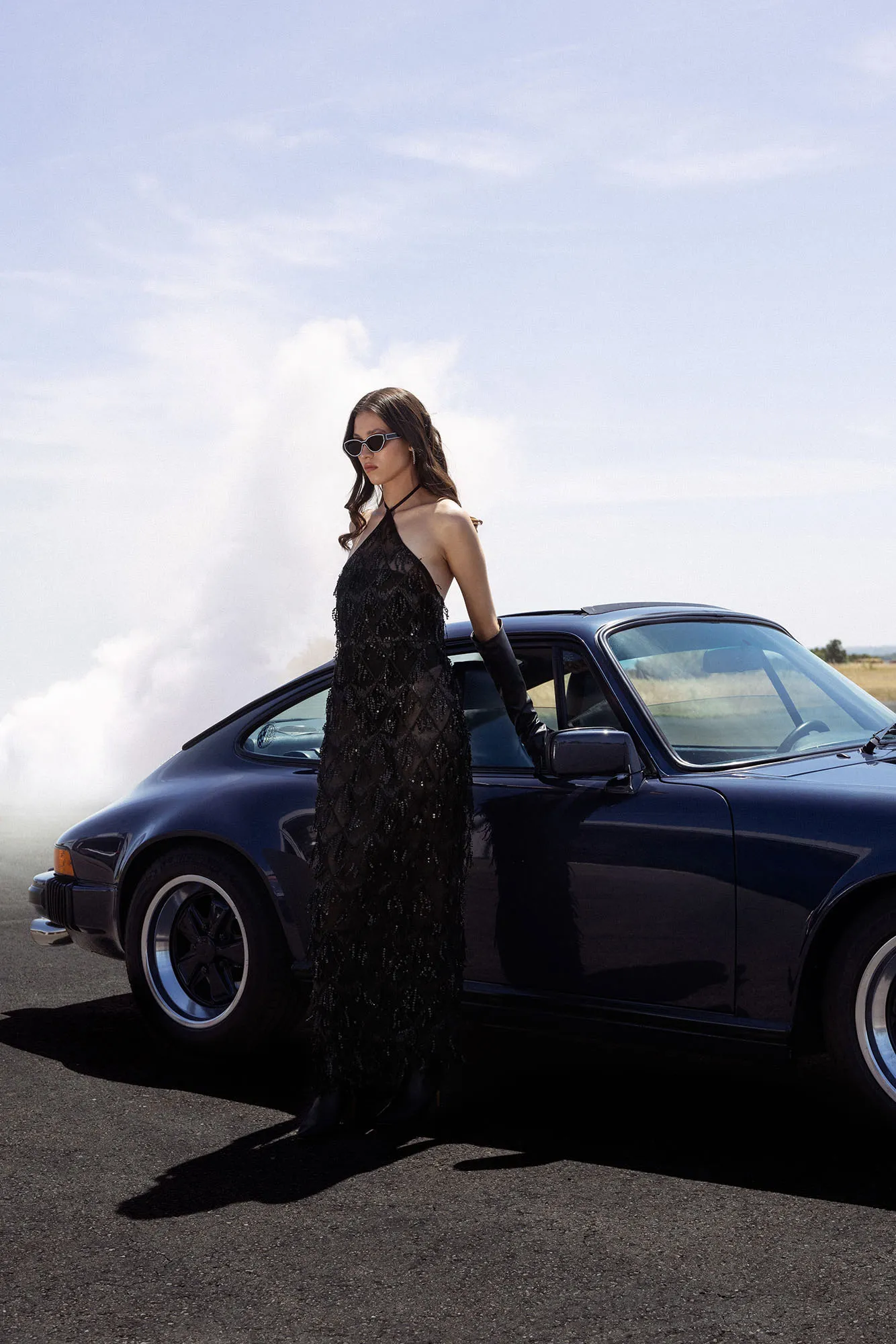 Shooting fashion et éditorial avec une Porsche 911 bleu foncé. Une mannequin en robe noire et lunettes de soleil pose devant la voiture.