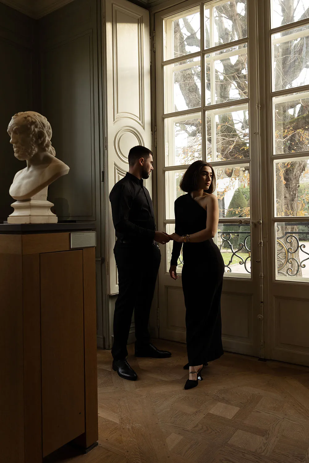Shooting photo de couple minimaliste et intimiste dans un musée parisien, jouant avec l'ombre et la lumière à la fenêtre et une statue en premier plan.