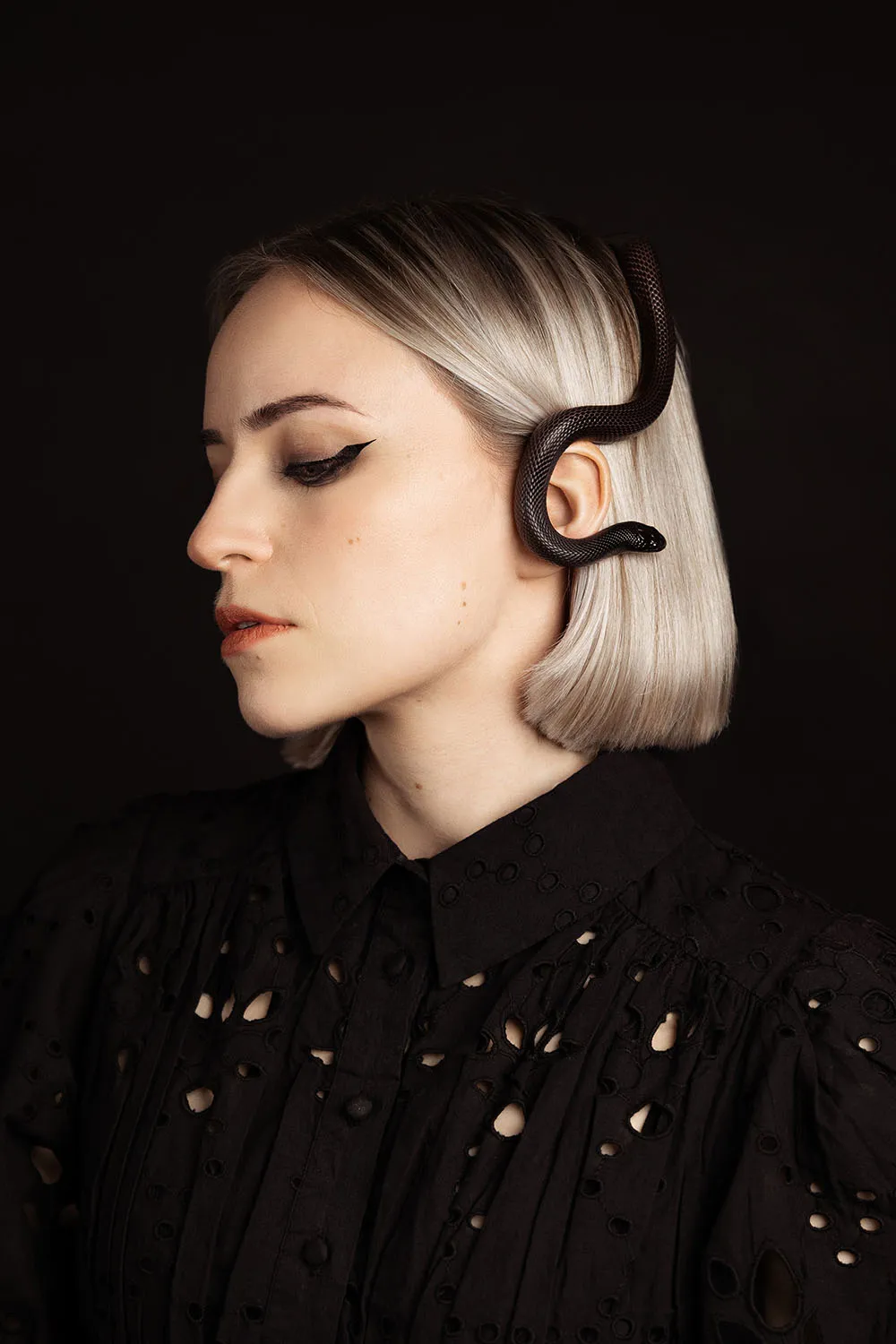 Photographie de portrait artistique en studio sur fond noir, modèle avec un petit serpent noir dans ses cheveux blonds.