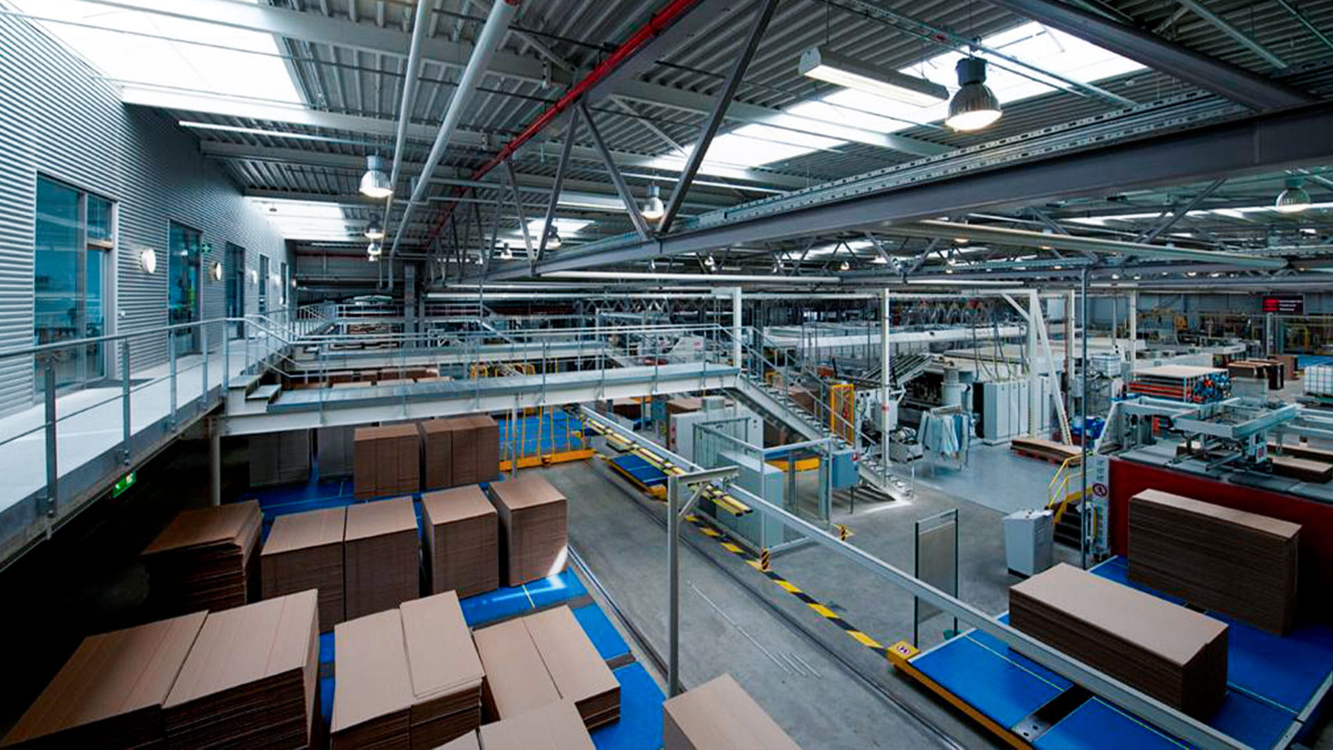 Interior of a large industrial warehouse with stacked cardboard boxes and conveyor belts under a metal roof structure
