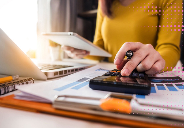 A person in a yellow shirt calculates with a calculator, holding a tablet. Papers with charts, a laptop, and clipboard are on the desk, conveying a busy work environment.