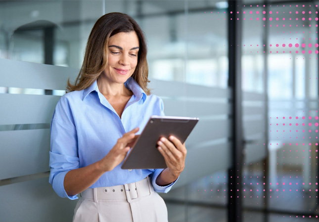 Smiling woman in a blue shirt uses a tablet, standing in a modern office with glass walls.