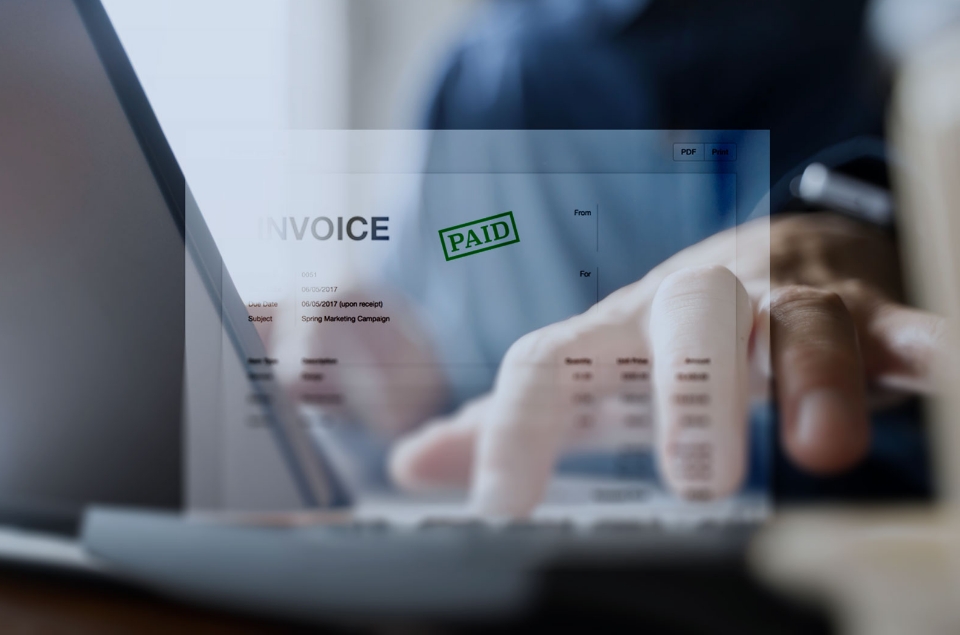 A close up of a person's hand typing on a laptop keyboard with an invoice on the screen marked as "PAID."